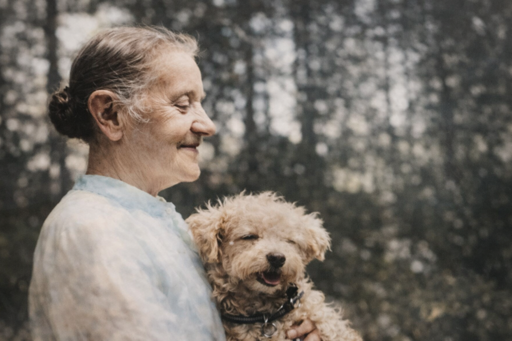 Portrait of an elderly woman with gray hair in a bun holding a small curly tan dog outdoors, both softly lit against a blurred forest background.