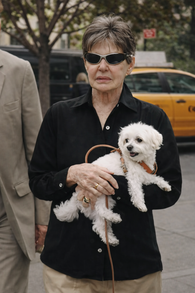 Eleanor Helmsley wearing dark sunglasses and a black blouse, holding a small white dog on a leash, standing on a city sidewalk with a yellow taxi and urban signage in the background.