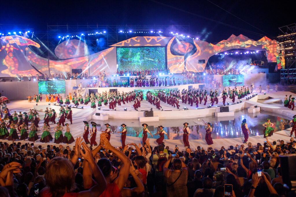 Wide view of a large outdoor nighttime performance featuring dozens of dancers in colorful costumes on a multi-tiered stage with bright lights and oversized decorative backdrops, while a packed audience in the foreground watches and records on their phones beside a reflective pool.
