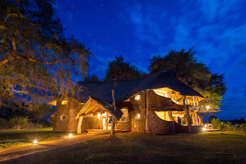 A warmly lit thatched-roof safari lodge sits beneath a star-filled night sky, surrounded by trees and illuminated pathways across the grassy grounds.