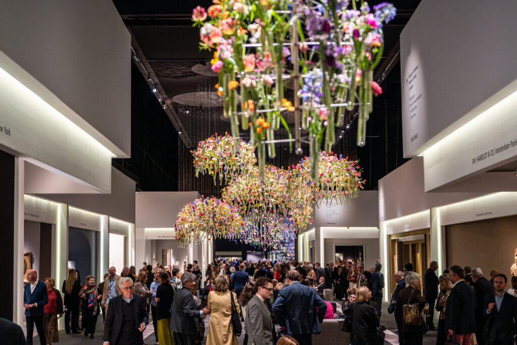 Crowded gallery hall at TEFAF Maastricht with visitors viewing art booths beneath large suspended floral installations made of colorful flowers hanging from the ceiling.