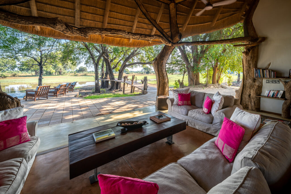 Open-air safari lodge lounge with cream sofas and bright pink cushions arranged around a wooden coffee table, overlooking a shaded deck with chairs beside a river and trees.