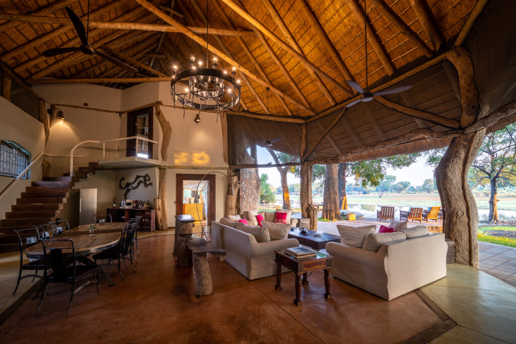 Open-air safari lodge interior with a high thatched ceiling, rustic wood beams, and a chandelier above a living area with white sofas, overlooking a riverside landscape through large open walls.