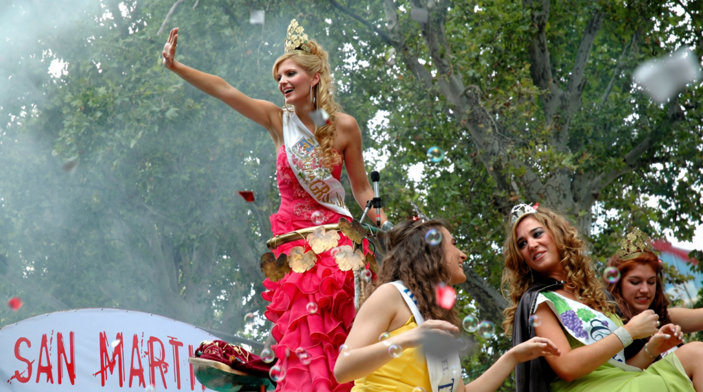 A crowned festival queen in a bright pink ruffled gown and sash waves from a parade float during a celebration, surrounded by other young women wearing tiaras and sashes, with confetti and trees in the background.