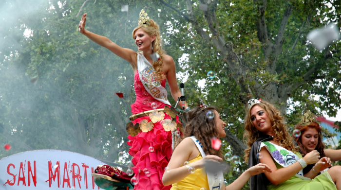 A crowned festival queen in a bright pink ruffled gown and sash waves from a parade float during a celebration, surrounded by other young women wearing tiaras and sashes, with confetti and trees in the background.