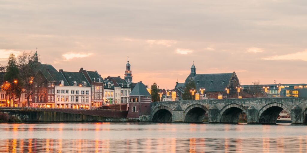 A calm riverside cityscape at sunset with warm lights glowing from historic buildings. A stone arch bridge spans the water, reflecting soft orange and pink tones from the sky, while church towers and traditional European architecture rise in the background