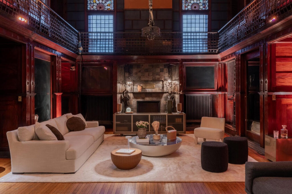 A grand lounge inside the Armory on Park Avenue in New York, featuring dark wood paneling, stained-glass windows, and a mezzanine balcony, styled with contemporary cream sofas, modern accent chairs, a round coffee table, and warm ambient lighting around a central fireplace.