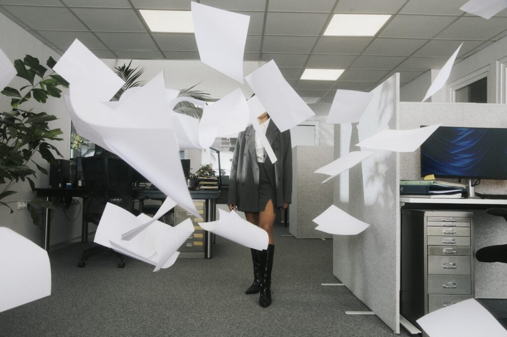 Woman in an office walking through cubicles as printer paper flies around her.