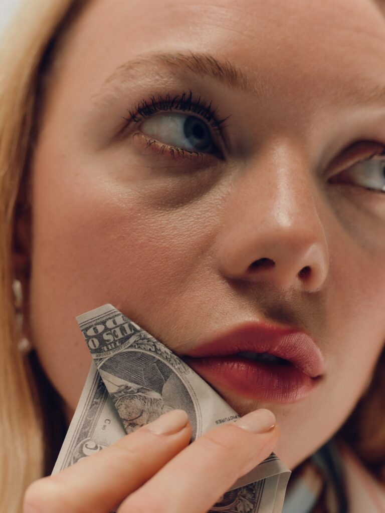 Close up of a woman in red lipstick holding a dollar to her lips and looking away from the camera.