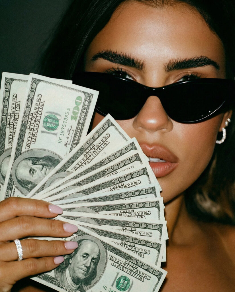 Close up on a woman holding a fanned-out stack of dollar bills and wearing black sunglasses.