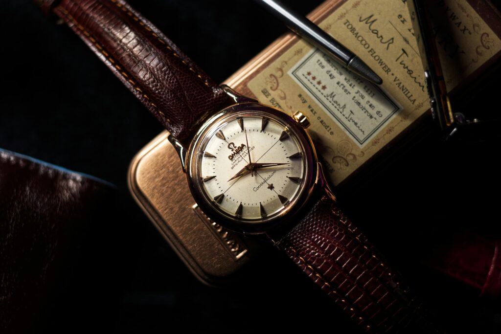 Vintage Omega Constellation wristwatch with a cream dial and gold case on a brown leather strap, styled beside a fountain pen and a tobacco-scented candle box in warm, moody lighting.