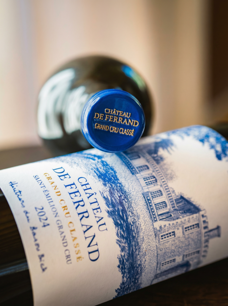 Close-up of a Château de Ferrand Saint-Émilion Grand Cru wine bottle lying on its side, with the blue capsule and illustrated blue-and-white label featuring the château prominently in focus against a softly blurred background.