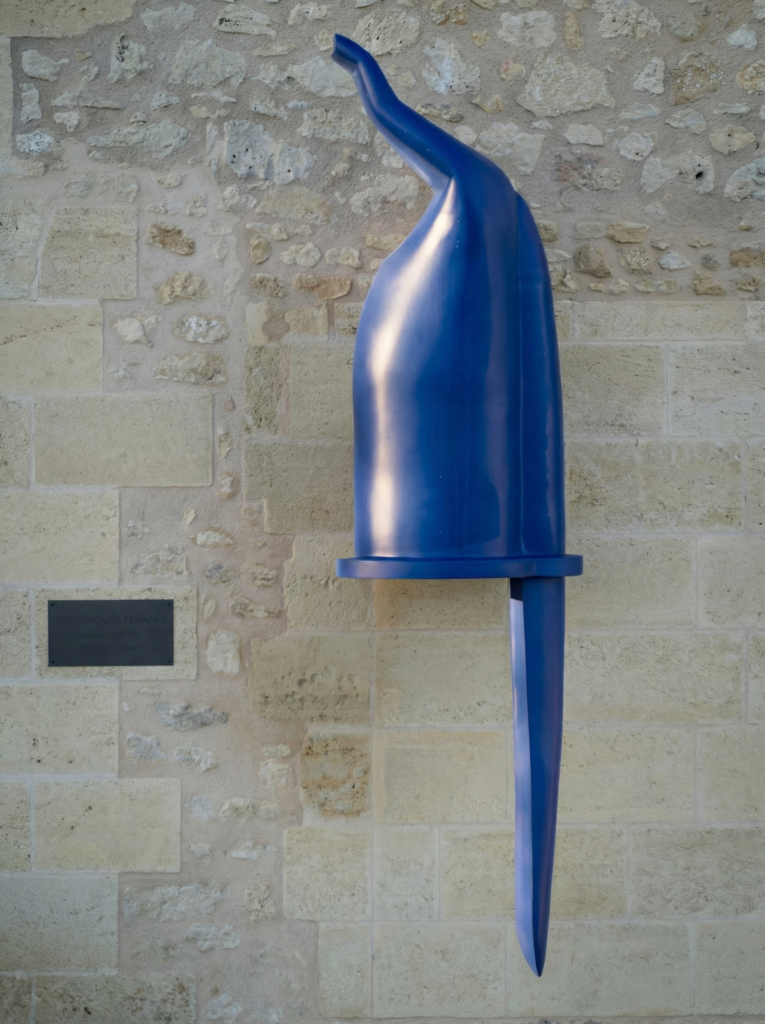 A tall, smooth, abstract blue sculpture with a curved top and pointed bottom is mounted on a light stone wall, with a small dark rectangular plaque positioned to its left.