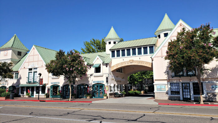 Wide street-level view of a charming, European-style shopping courtyard with cream-colored buildings, green metal roofs, and turreted towers. An arched passageway connects the structures, with small storefronts beneath green awnings, flowering trees along the sidewalk, and a clear blue sky overhead.