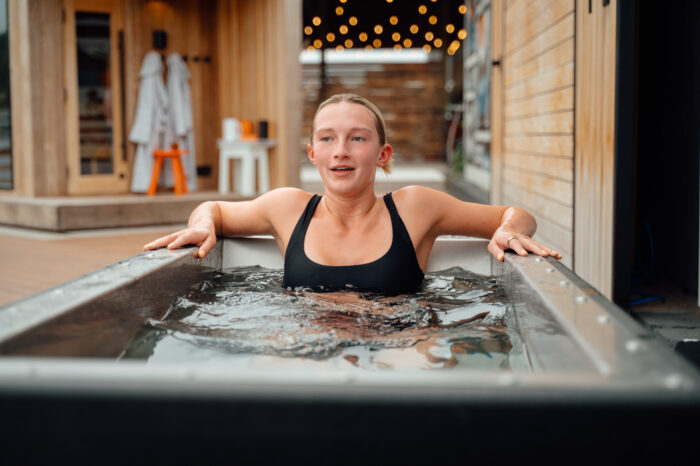 A woman in a black swimsuit sits immersed in a cold plunge tub, gripping the edges as water ripples around her. She appears focused and slightly tense from the cold. Behind her, a warm, wood-clad outdoor setting includes a sauna entrance, hanging robes, small stools, and soft string lights creating a cozy contrast to the icy water.
