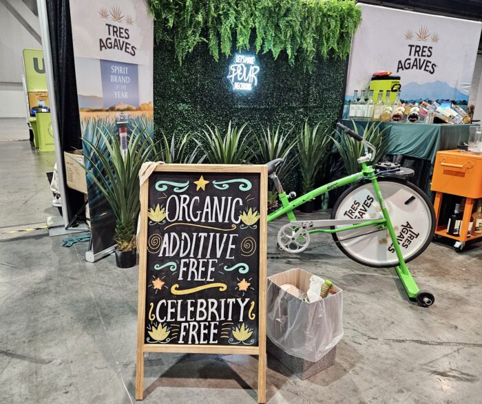 Trade show booth for Tres Agaves featuring agave plants, a neon sign, tequila bottles, and a chalkboard reading “Organic, Additive Free, Celebrity Free,” with a green branded bicycle display.