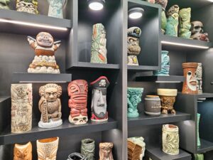 Shelves displaying a colorful collection of carved tiki mugs and decorative ceramic vessels, featuring detailed faces, figures, and tropical motifs under soft lighting.
