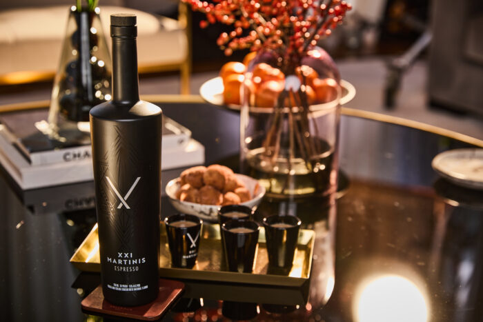 Bottle of XXI Martinis Espresso with branded shot glasses on a tray, set on a glossy table with cocktails, snacks, and warm ambient lighting in a lounge setting.