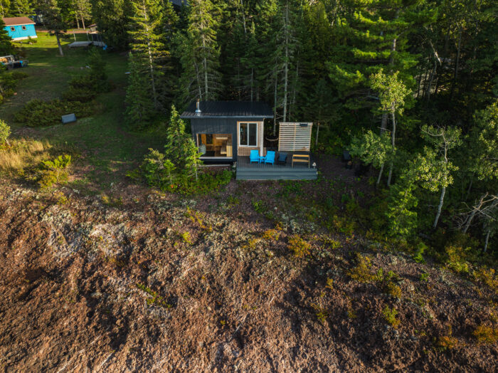 Aerial view of a small modern cabin nestled among tall trees on a rocky shoreline. The cabin features a dark exterior, large windows, and a compact deck with bright blue chairs. Surrounding the structure are dense evergreens and a few nearby buildings set back in a clearing, with the rugged terrain sloping down toward the foreground.