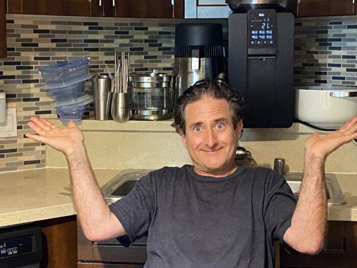 Corey Levitan, seated in his kitchen, raises both hands with a playful, exasperated expression, surrounded by stainless steel containers and appliances, with a stack of silicone food storage bags floating beside him.