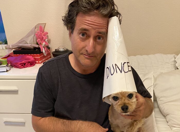 Man holding a small tan dog wearing a paper cone labeled “DUNCE,” both looking at the camera indoors.