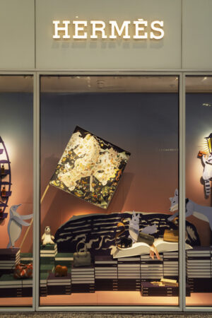 Hermès storefront window display featuring artfully arranged handbags, books, and whimsical animal illustrations beneath a glowing Hermès sign.

