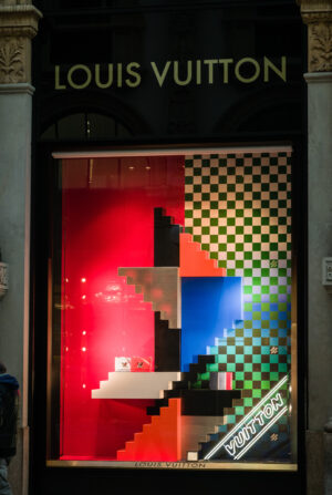 Louis Vuitton storefront window display featuring bold geometric stair-like structures in red, blue, and green with checkered patterns, showcasing handbags beneath the brand’s illuminated logo.
