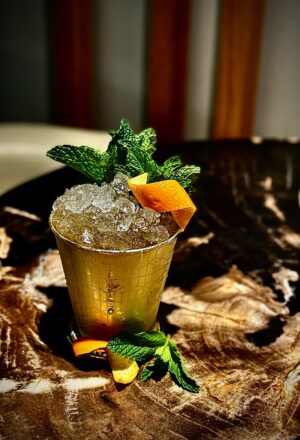 A gold-toned cup filled with crushed ice holds a Blackwood Julep cocktail garnished with fresh mint and an orange peel, set on a richly grained wooden surface with warm, ambient lighting.