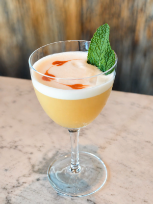 A pale yellow cocktail in a stemmed coupe glass, topped with a smooth foam and streaks of orange-red bitters, garnished with a fresh mint leaf, set on a light marble surface with a softly blurred wood background.