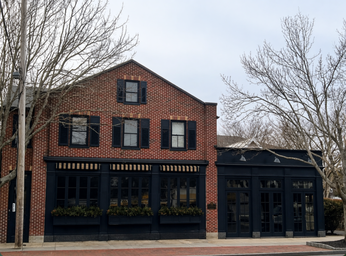 Exterior view of East Hampton Grill, a two-story red brick building with black shutters and trim, featuring large front windows, striped awnings, and window box planters. Bare trees frame the scene along a quiet street, giving it a refined, understated Hamptons feel in the off-season.