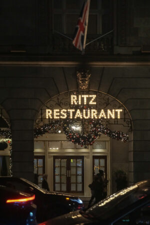 Illuminated “Ritz Restaurant” sign above an ornate entrance at night, framed by holiday garlands and a Union Jack flag, with cars and pedestrians passing below.
