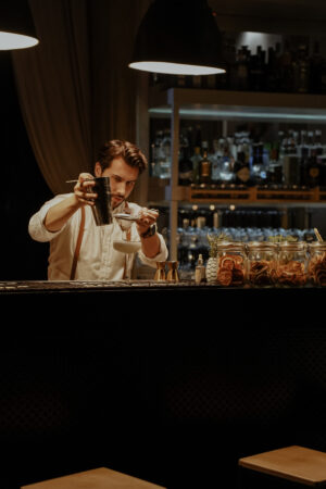 A bartender in a dimly lit, intimate bar carefully strains a cocktail into a coupe glass, focused under warm pendant lighting, with shelves of spirits and jars of dried citrus softly glowing in the background, creating a mood of precision, craft, and quiet intensity.
