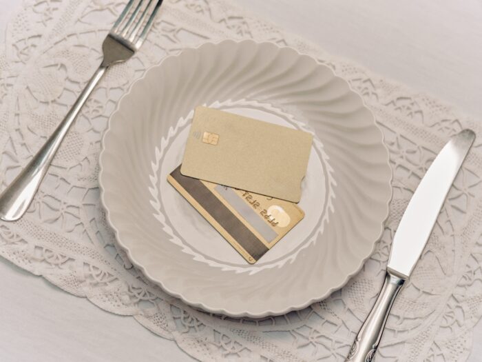 A white, scalloped-edge bowl set on a lace tablecloth holds two gold credit cards instead of food, with a fork and knife placed on either side, creating a minimalist, symbolic dining scene about consumption and wealth.