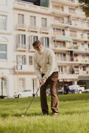 An older man in a cream knit sweater, brown trousers, and a dark flat cap addresses a golf shot on a grass median in an urban setting, with a European residential facade softly blurred behind him.