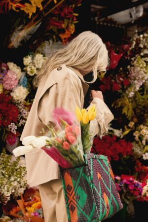 A person with long light-blonde hair in a beige trench coat browses a vibrant outdoor flower stand, carrying a colorful woven bag filled with tulips.