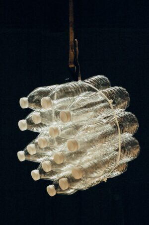 Alt text: A cluster of empty plastic water bottles bound together with zip ties hangs from a strap against a dark background, creating a sculptural arrangement that highlights plastic waste and reuse.