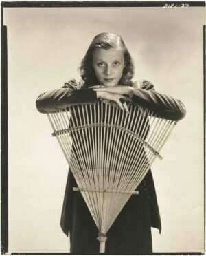 Black-and-white portrait of Eleanor Lambert as a young woman, resting her chin on her hands atop a fan-shaped wooden prop, gazing directly at the camera with a poised, confident expression.