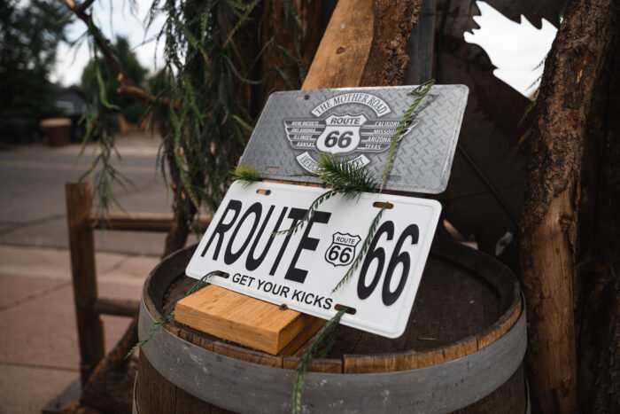 Close-up of two vintage-style Route 66 signs displayed on a wooden barrel. One sign reads “Route 66 – Get Your Kicks,” while the other features “The Mother Road” with listed states. Pine branches and rustic wood elements surround the signs, with a softly blurred street scene in the background.
