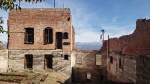 Partially collapsed brick building with exposed interior walls and empty window frames, revealing weathered wood and peeling plaster. The structure sits on a hillside with expansive desert and canyon views in the distance, under a clear blue sky, evoking a historic, abandoned mining-town atmosphere.
