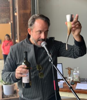 John Heers, co-founder and director of First Things Foundation, speaks into a microphone while holding a bottle of wine in one hand and raising a traditional horn-shaped drinking vessel in the other, addressing an indoor gathering with a casual, intimate atmosphere.