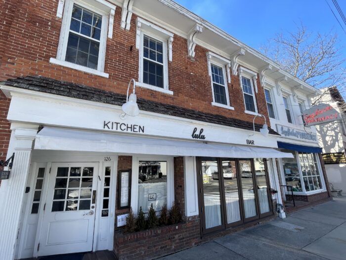 Street-level view of a classic Sag Harbor storefront featuring Lulu Kitchen & Bar, with a red brick facade, white awning, and simple black lettering. Large front windows and glass doors line the entrance, while traditional white trim and gooseneck light fixtures add to the understated Hamptons charm.