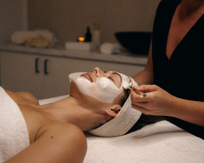 Client reclines during a spa facial as an esthetician applies a white treatment mask with a brush in a softly lit, serene setting.
