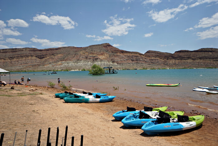 Sunny lakeside scene at Quail Creek, with colorful kayaks pulled up along a sandy shore. People wade and swim in the calm water, while paddleboards and small boats dot the lake. In the distance, layered desert hills rise beneath a bright blue sky with scattered clouds.
