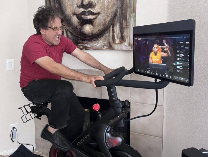 The author rides a Peloton stationary bike in a home setting, leaning forward with intensity as a virtual instructor leads a class on the screen; behind him, a large Buddha artwork hangs above a fireplace, contrasting the calm decor with the energy of the workout.