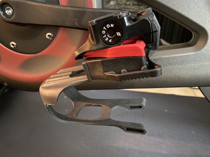 Close-up of a Peloton bike pedal and cleat mechanism, showing the black pedal with a red clip-in system and adjustment dial, set against the matte frame of the stationary bike.
