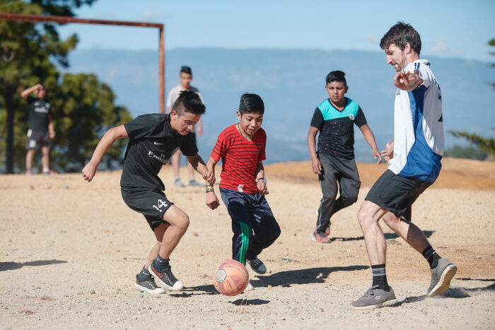 A group of boys play soccer on a dusty outdoor field under a clear blue sky, focusing intently on the ball as an adult joins in, guiding the play; a simple goalpost stands in the background with trees and distant hills beyond.