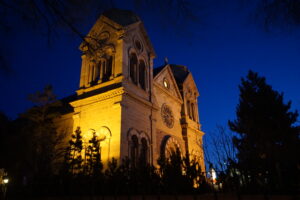 Low-angle nighttime view of a historic cathedral illuminated in warm golden light, highlighting its stone façade, arched entryways, and twin bell towers. A circular rose window sits above the main entrance, while dark tree silhouettes frame the building against a deep blue evening sky.