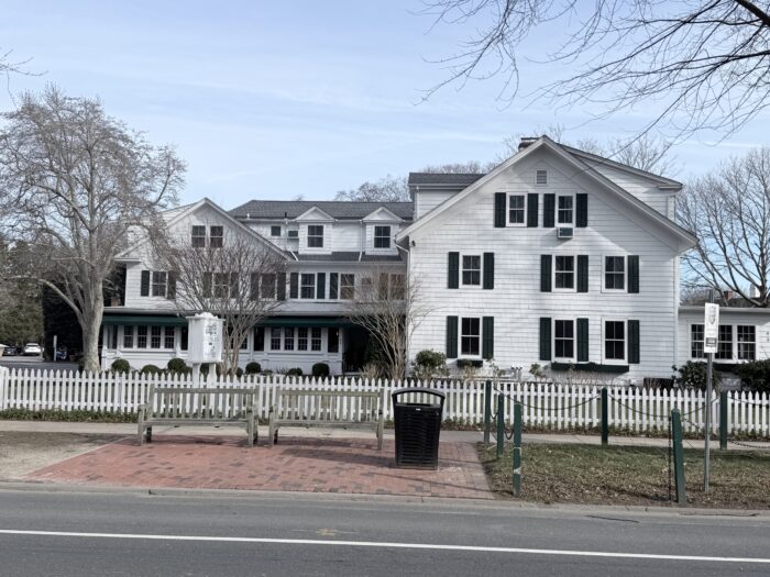 Exterior view of a classic Hamptons inn-style building with white clapboard siding and dark green shutters, set behind a white picket fence along a quiet street. Bare trees and a brick sidewalk frame the scene, with a sign marking The Palm at the Huntting Inn, conveying a timeless, understated East Hampton setting.