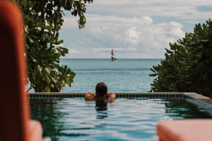 Person relaxes in an infinity pool overlooking a calm ocean, framed by lush greenery, with a sailboat on the horizon under a cloudy sky.