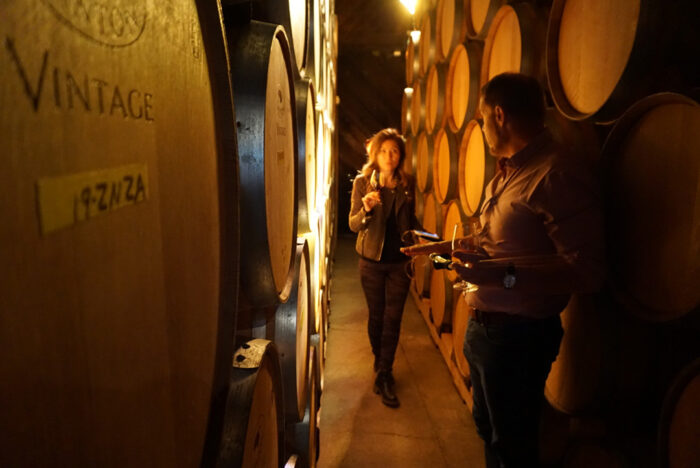Two people walking through a dimly lit wine cellar lined with oak barrels, holding glasses of wine.
